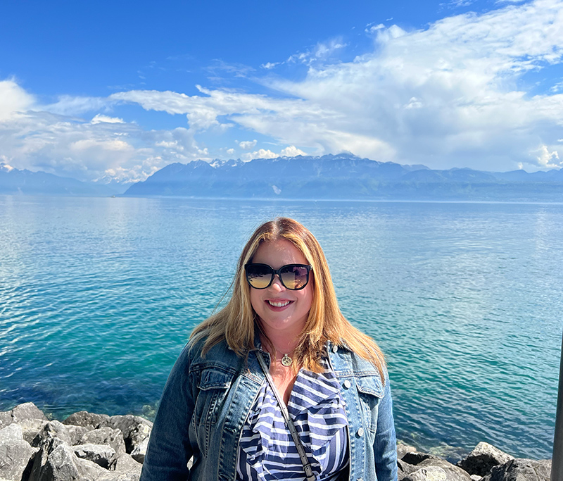 Dezaree standing on rocks in front of a body of water with mountains and clouds in the distance