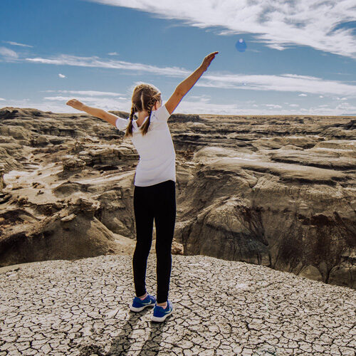girl standing on a mesa with her hands in the air and blue skies above